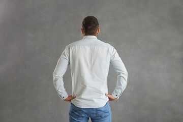 View from behind of athletic broad shouldered European man standing hands on hips against grey background. Studio backside shot of young guy with short hair in light blue denim shirt with mock up back - Powered by Adobe