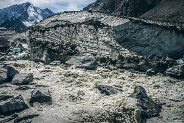 Scenic landscape with powerful mountain river beginning from glacier among large moraines on background of great snowy mountains. Beautiful scenery with glacier at source of turbulent mountain river.