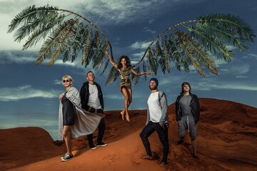 Groupe of fashion people are on red dunes, in center young female angel with palm wings