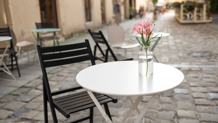 Fototapeta na wymiar Beautiful delicate pink flower in a transparent glass vase on a white street table. Street view with cafe terrace in the morning in the city