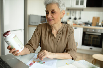 Pretty grey haired mature stylish woman with earrings doing online shopping, reading information on vitamin complex bottle at kitchen table, ready to make new order on website of dietary supplements