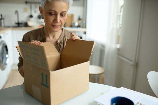 Crop shot of senior Caucasian woman wearing stylish haircut and make-up, unpacking delivery box of her online order at home, looking inside with interest, dressed in lovely brown blouse