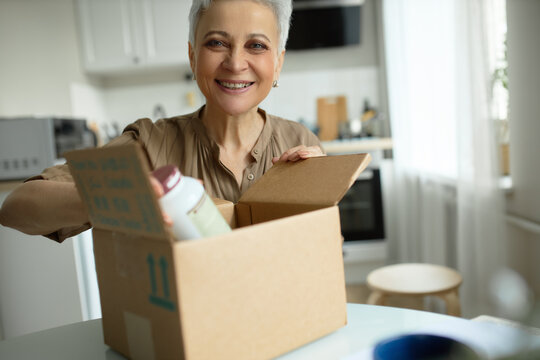 Crop shot of cheerful elderly female with makeup and manicured nails, dressed casually, opening package with organic dietary supplements. Happy and satisfied with fast delivery services