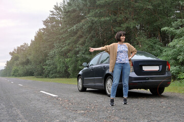 A young woman standing near a broken car with a raised hood is experiencing problems with her...