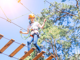 Kid in orange helmet climbing in trees on forest adventure park. Girl walk on rope cables and high suspension bridge in adventure summer city park. Extreme sport equipment helmet and carabiner.