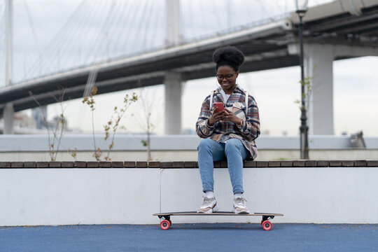 Happy african american skateboarder girl reading message with good news on smartphone screen sitting on longboard in modern urban space over bridge and smiling. Technology and communication concept