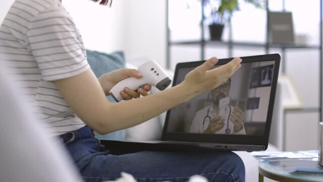 Close up of unrecognizable woman, sitting on sofa at home, measuring her body temperature with non-contact infrared thermometer under supervision of male doctor by video call. Blur view of laptop