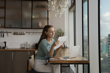 Happy young woman reading paper letter at home workplace with laptop, getting good news, bank or...