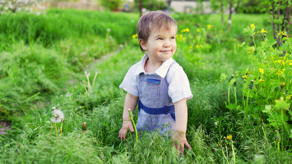 Cute boy in fashionable clothes with blue eyes plays and jumps in the tall grass in a large green blooming garden