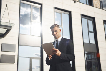 Confident mature businessman holding digital tablet while standing near office building