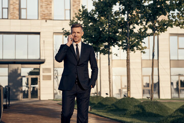 Confident mature businessman talking on mobile phone while walking near office building outdoors