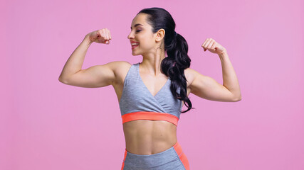 Fototapeta na wymiar cheerful sportswoman showing muscles isolated on pink