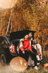 A man and a woman in love in warm red sweaters travel together relax on a picnic in a romantic adventure traveling by car in an fall forest in nature among trees and foliage in autumn outdoors