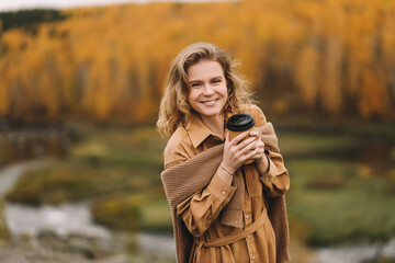 A happy pretty smiling young woman in casual warm clothes enjoys solitude drinking coffee walking traveling by car in the autumn forest in nature golden fall