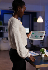 Concentrated african american executive manager holding tablet computer in hands looking at company profit planning financial strategy. Overworked businesswoman working late at night in startup office