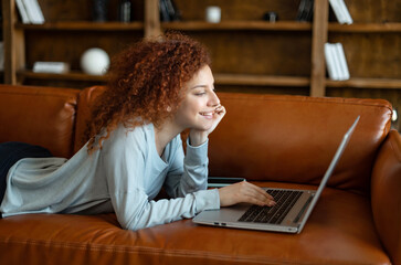 Cheerful curly red-haired woman spends leisure online, smiling woman lying on the couch with a laptop, watching movies, scrolling news feed in social media, looks with interest at screen and laughs