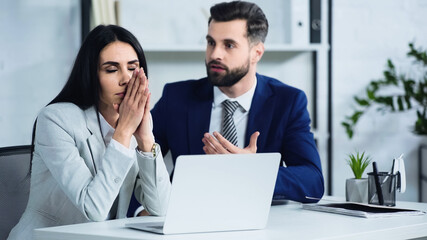 upset businesswoman with closed eyes near blurred and displeased businessman quarrelling in office