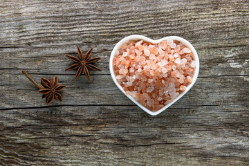 A porcelain bowl in the shape of a heart filled with large crystals of pink himalayan salt on an old weathered board, next to it there are star anise inflorescences. 