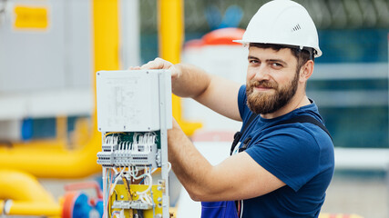 Electrician in a helmet and overalls works adjusts the equipment looks into the camera and smiles....