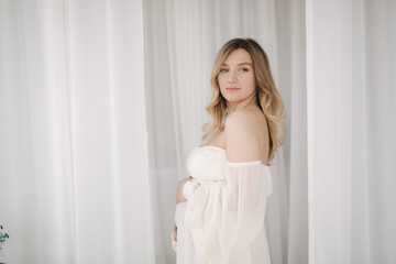 Fototapeta na wymiar Beautiful pregnant woman in elegant white dress posing to photographer in studio. Background of white tulle