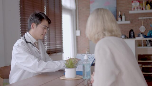 Senior Caucasian woman consults with young Asian doctor. Doctor is describing health checking result to patient during home visit  medium close-up with slide movement.  