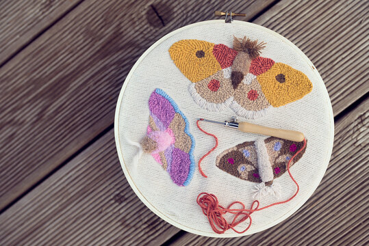 Top view of three embroidered multicolored butterflies on two-thread cloth in a hoop with punch needle and brick-red thread inside of it. Punch needle embroidery project on wooden panels.