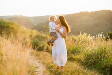 Joyful mother with her little son in her arms. Elegant woman with little boy walking in the field with beautiful landscape