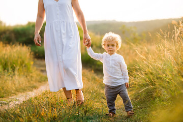 Naklejka na ściany i meble Beautiful woman in light white dress leads her son by the hand along the path of a picturesque field