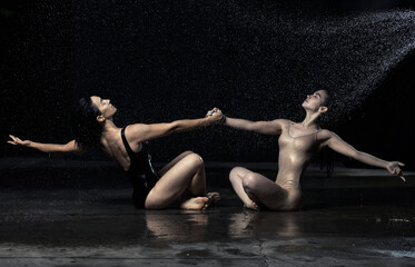 two beautiful women of Caucasian appearance with dark hair are sitting on the asphalt in drops of water on a black background