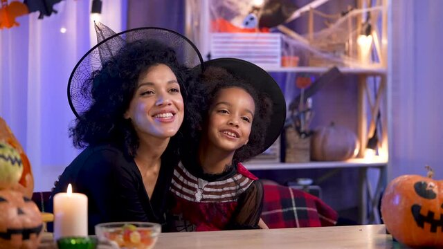 Mom and daughter African American in festive costume and witch hat have fun and hug joyfully. Happy family sits at table in room decorated for the Halloween night. Close up. Slow motion.