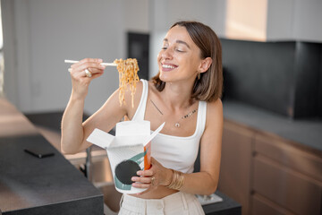 Young woman eating noodles with chopsticks from cardboard packaging in the kitchen at home....