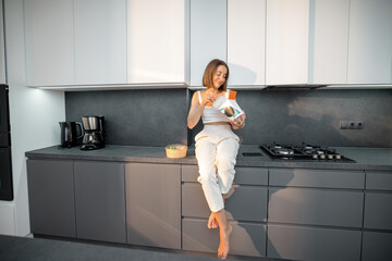 Young woman eats takeaway food while sitting on the modern kitchen. Concept of ordering food and not cooking at home. Wide view on the modern kitchen interior