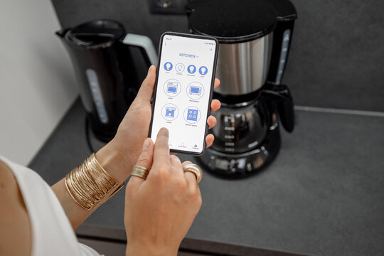 Smart Phone With Running Mobile Application To Control Smart Devices In The Kitchen. Female Controlling Coffee Machine Or Electric Teapot With Phone At Home. Smart Home Concept