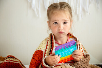 A sad girl sits in a knitted blanket wrapped up and holds a pop game in her hands. The concept of the game at home.