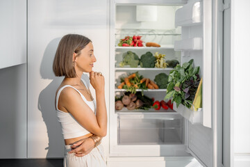 Young woman standing near the fridge full of fresh vegetables at modern kitchen. Healthy vegan eating concept