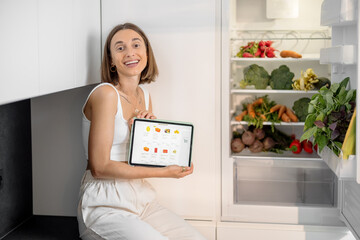 Woman sitting near the fridge full of fresh vegetables, holding digital tablet with running e-shop...