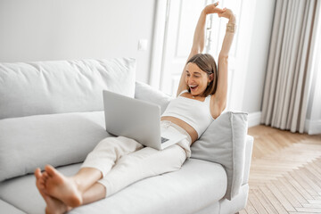 Fototapeta premium Young woman stretching up, having a break while working on laptop on the couch at home. Coziness and work from home concept