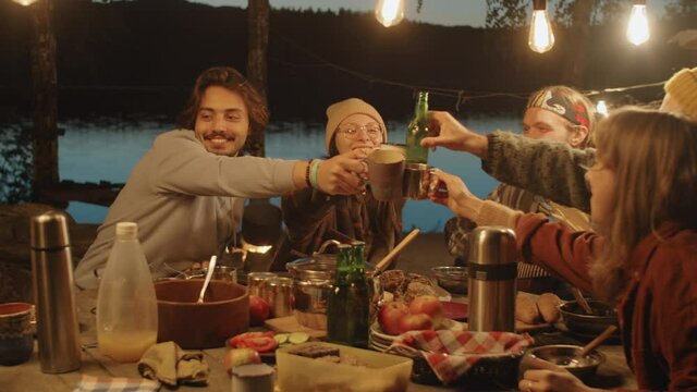 Young cheerful friends clinking cups in toast while man playing guitar at dinner table at campsite in evening