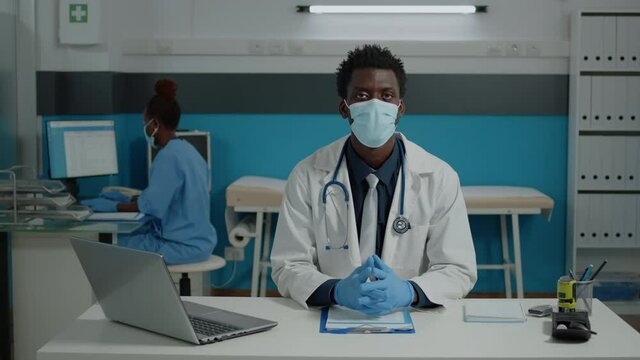 Portrait of african american doctor looking at camera wearing white coat in office at medical facility. Black medic sitting at desk with laptop, tools, stethoscope, and documents