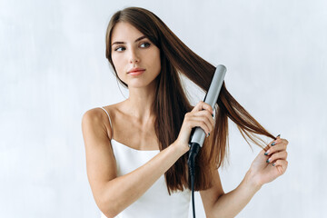 Closeup portrait of a smiling young woman in domestic clothes using a hair straightener while...