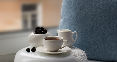 A white porcelain cup with black coffee on a saucer, a creamer, a vase with blackberries and a lace ribbon on a glass stand against the background of a decorative pillow and a window.
