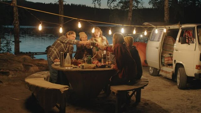 Group of young cheerful friends clinking drinks in toast while having dinner at campsite by lake in evening