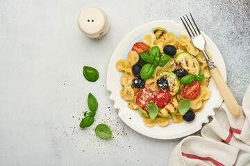 Pasta fiocchi salad with vegetables grilled zucchini, cherry tomato, olives, basil and parmesan cheese in white plate on light slate, stone or concrete background. Lunch concept. Top view
