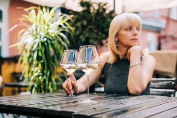 Thoughtful adult mature woman sitting in bar outdoors with wine glasses and blurry restaurant background scene, drinking white wine. Summer sunny day on patio. People lifestyle