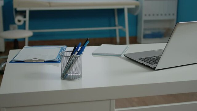Close up of modern desk in empty doctors office at medical clinic with laptop, document files, notebook and pens. Nobody in healthcare checkup room designed with professional instruments