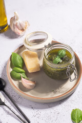 A small mason jar with green italian basil sauce pesto on a grey plate with hard cheese pecorino, garlic cloves and fresh basil leaves