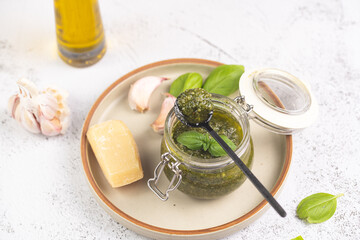 A small mason jar with green italian basil sauce pesto on a grey plate with hard cheese pecorino, garlic cloves and fresh basil leaves
