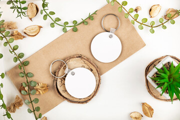 From above modern keychain mock ups with blank round pendants placed on piece of wood and rectangular cardboard near green plants and dried leaves on white background. Key chain mock up - obrazy, fototapety, plakaty
