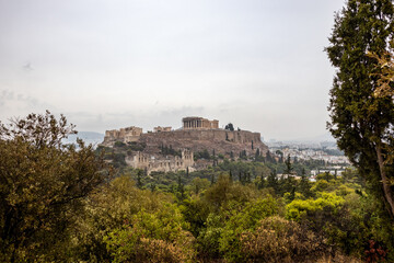 Fototapeta na wymiar Acropolis hill (Parthenon, Propylaea, Temples, Odeon of Herodes Atticus) view through summer greenery. Athens ancient historical landmark in city center from Filopappou Hill on cloudy day