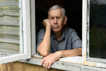 Portrait of a sad elderly man of 80 years old, sitting in the window of an old wooden house. 